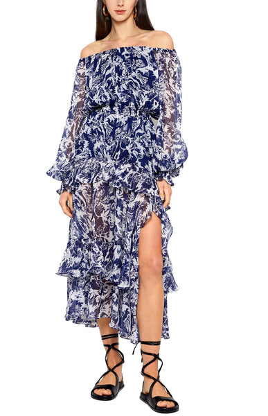 Woman in a blue and white floral Prabal Gurung off-the-shoulder ruffle dress with black strappy sandals.