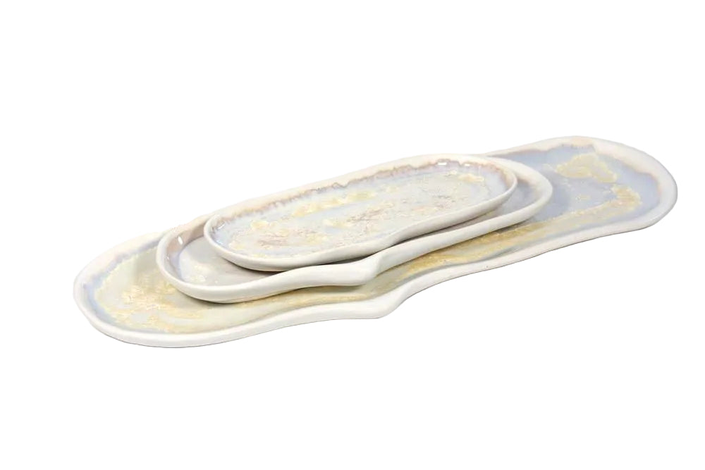 The AE Ceramics Large Razor Clam Tray Collection features three stackable ceramic trays with irregular, coastal-inspired shapes and glazed light-toned surfaces accented by subtle decorative patterns.