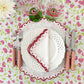 A table setting with white plates, a scalloped napkin, the Duggan Society Rattan Pink 5 Piece Flatware Set, two glasses, and a floral tablecloth in pink and green.