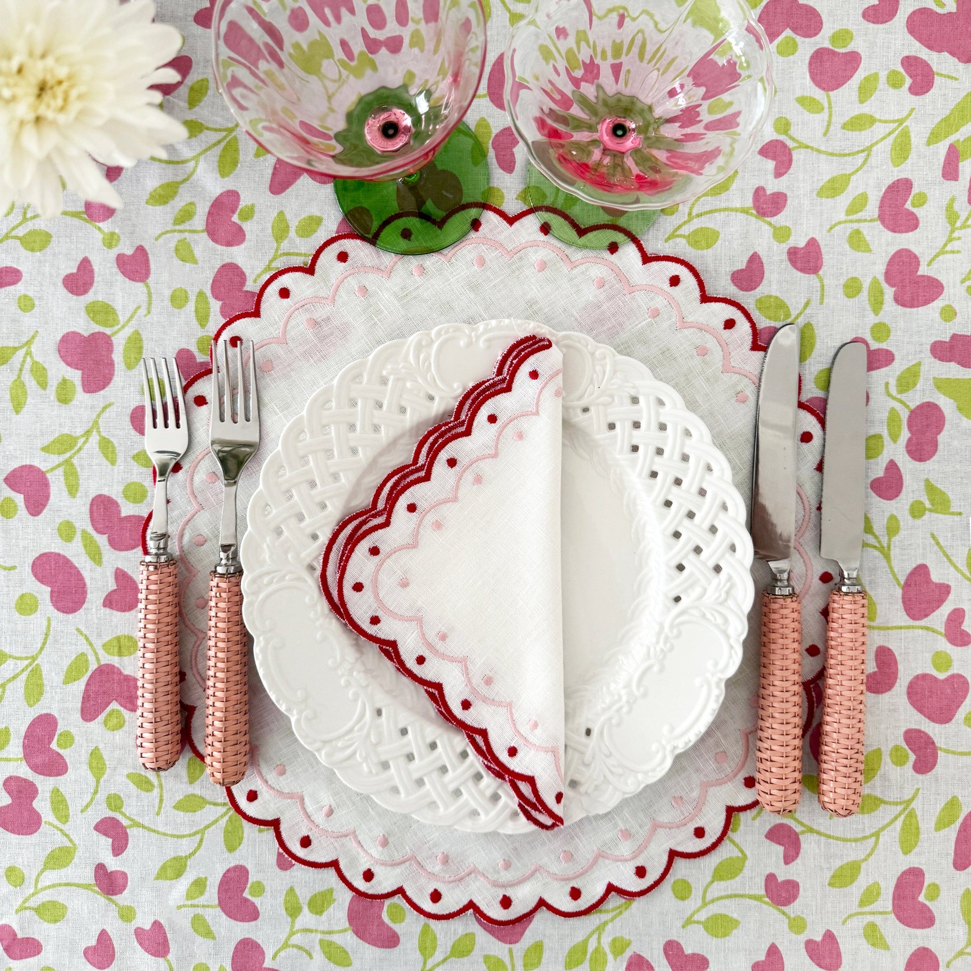 A table setting with white plates, a scalloped napkin, the Duggan Society Rattan Pink 5 Piece Flatware Set, two glasses, and a floral tablecloth in pink and green.