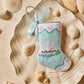 The Palm Yacht Needle Point Stocking by Bauble Stockings, a small pastel nautical Christmas ornament with a blue ribbon, rests on a shell-shaped dish surrounded by assorted seashells.
