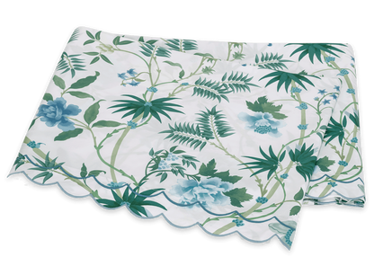 The Matouk Palmyra Bedding Collection, Emerald, features a folded tablecloth with green and blue palm fronds and leafy patterns on a white background, finished with a scalloped edge.