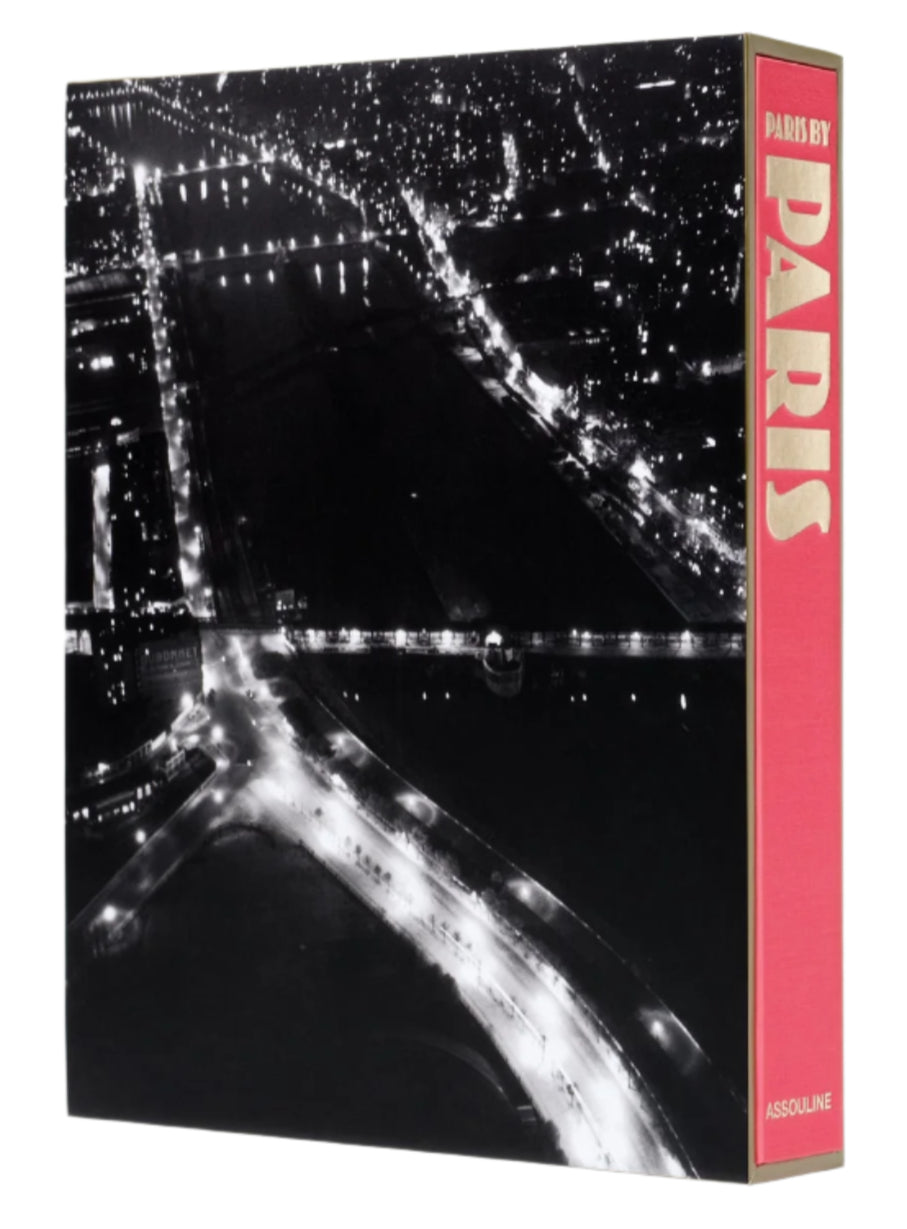 An aerial view of a cityscape at night shows illuminated roads and bridges, while the spine of "Paris by Paris" by Assouline, evoking Belle Époque charm, peeks out.