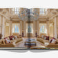 An open book, Passalacqua: A Love Letter to Lake Como by Assouline, displays a photo of an elegant living room with high ceilings, large windows, chandeliers, beige sofas, and ornate decor.