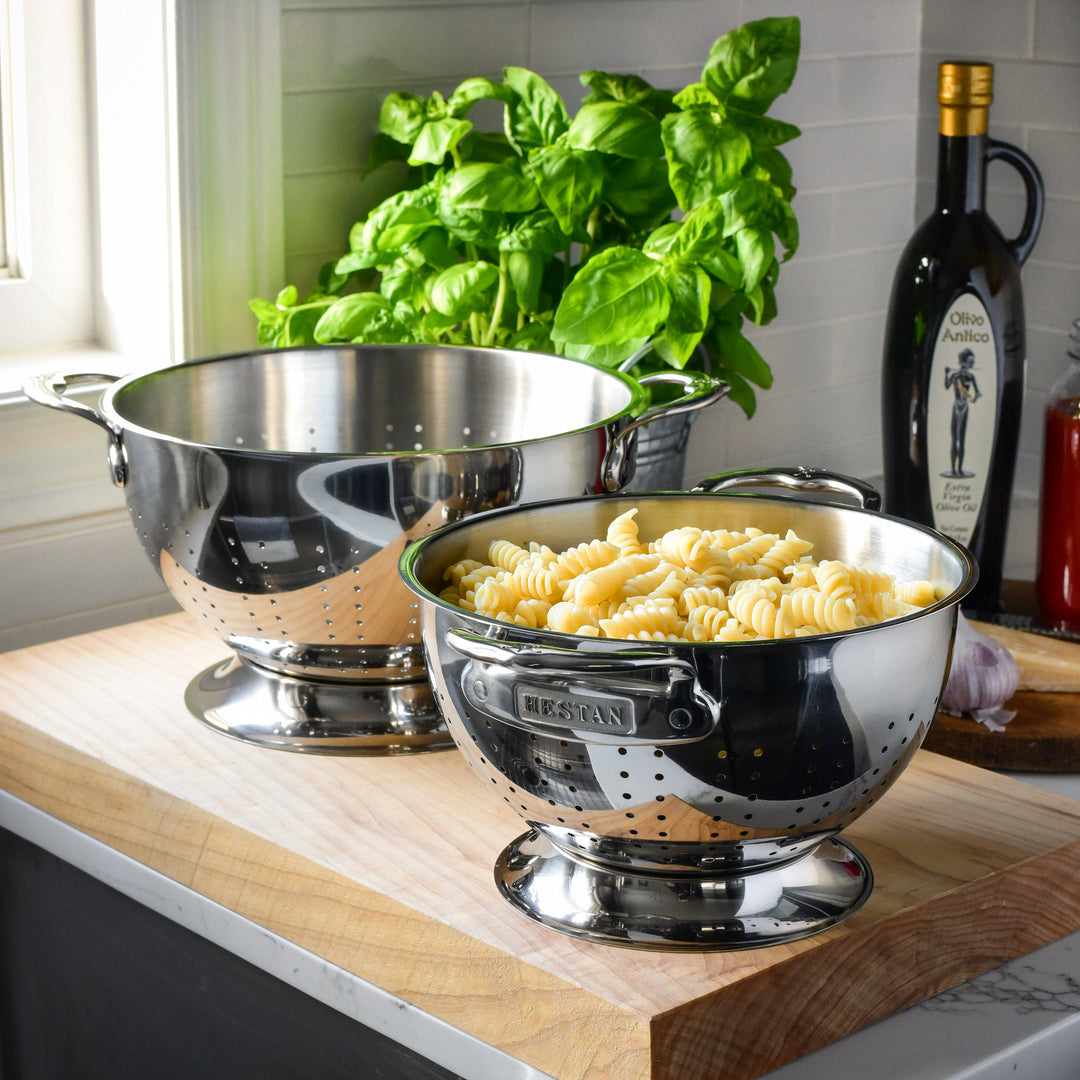 The Hestan Provisions Stainless Steel Colander 2-Piece Set features ergonomic handles and superior drainage; one colander holds cooked rotini while the other is empty, with fresh basil and olive oil in the background.