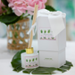 The Perfectly Palm Beach Signature Scent Collection reed diffuser, featuring a gold cap and branded label, sits by its matching box with a white ribbon on a green leaf, with pink flowers in the background.
