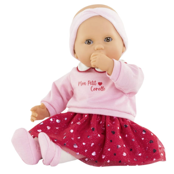 The Corolle Albane Heart Petals Doll from the Corolle collection wears a pink headband, a pink top with "Mon Petit Corolle" printed on it, and a red patterned skirt. This charming doll, made of soft vinyl, sits cutely with its thumb near its mouth.