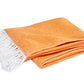 The Pezzo Throw Collection by Matouk features a folded orange cotton blanket with white fringe on one end, OEKO-TEX certified and made in Portugal, displayed on a white background.