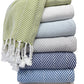 A stack of six folded textured towels in white, blue, and beige is topped with an A Soft Idea Nantucket Herringbone Throw Collection blanket featuring green-and-white stripes and hand-knotted fringe.