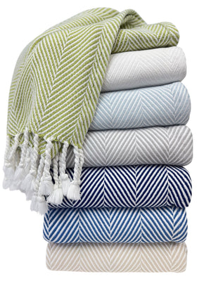 A stack of six folded textured towels in white, blue, and beige is topped with an A Soft Idea Nantucket Herringbone Throw Collection blanket featuring green-and-white stripes and hand-knotted fringe.