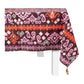 A rectangular table with a white base is draped in Figue’s Dancing Ikat Ganache Collection tablecloth, featuring purple, red, and pink geometric and floral motifs with a tassel accent on one corner.
