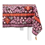 A rectangular table with a white base is draped in Figue’s Dancing Ikat Ganache Collection tablecloth, featuring purple, red, and pink geometric and floral motifs with a tassel accent on one corner.