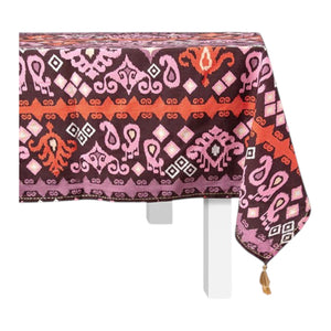A rectangular table with a white base is draped in Figue’s Dancing Ikat Ganache Collection tablecloth, featuring purple, red, and pink geometric and floral motifs with a tassel accent on one corner.