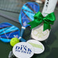 Haute Papier’s Letterpressed Pickleball Coasters—one stamped “I’LL DINK TO THAT”—are paired with two blue paddles and a yellow ball, all arranged on a pickleball court and finished with a green bow for a charming hostess gift.