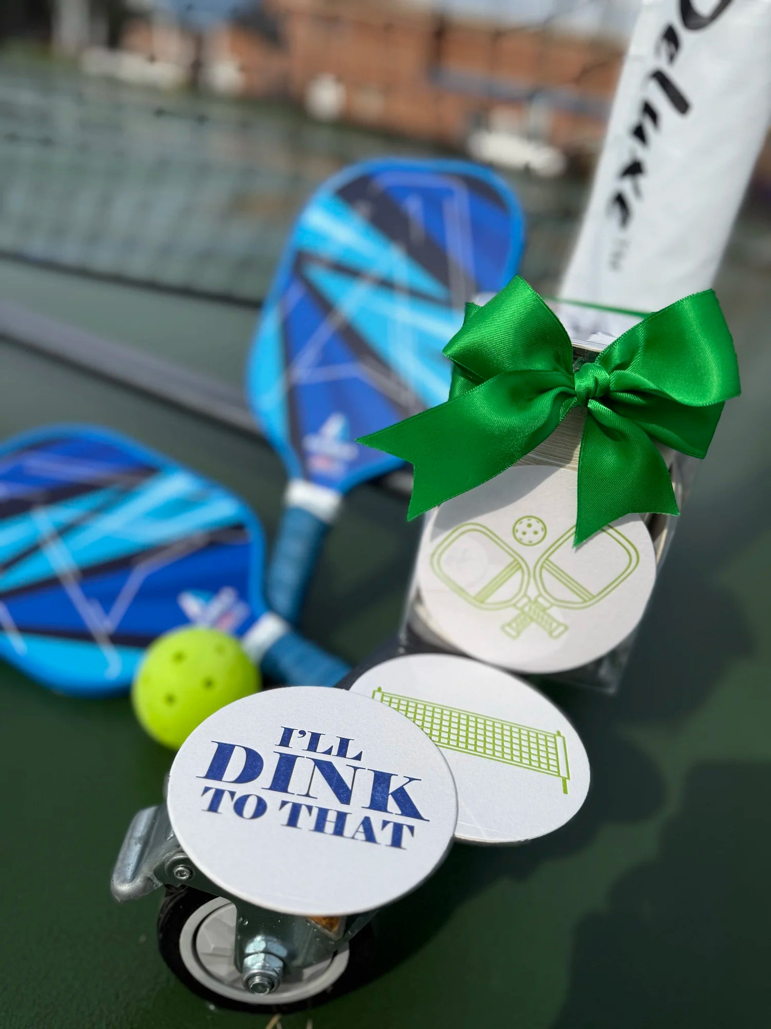 Haute Papier’s Letterpressed Pickleball Coasters—one stamped “I’LL DINK TO THAT”—are paired with two blue paddles and a yellow ball, all arranged on a pickleball court and finished with a green bow for a charming hostess gift.