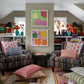 Two patterned armchairs with John Robshaw Kaavia Coral Decorative Pillows in a linen-cotton blend face each other in a room with bookshelves, flower vases, and two Jaipur block print–inspired floral artworks between windows.