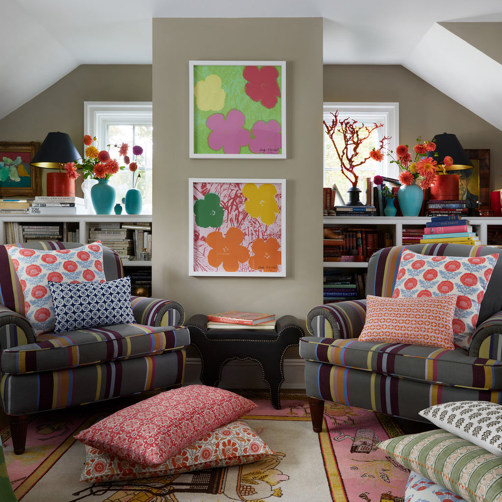 A cozy living room features two striped armchairs and the John Robshaw Yavi Coral Bolster, accompanied by floral cushions and colorful wall art. Bookshelves filled with treasures are enhanced by mirrors and vases of flowers, adding charm to the space.