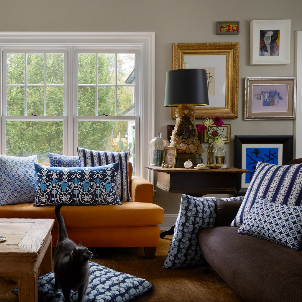 A living room with a yellow couch adorned with John Robshaw Mahin Indigo pillows, a brown sofa, a wooden table, and a black lamp. Various framed artworks decorate the walls as a black cat lounges comfortably on the floor.