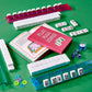 Graphic Image’s Play Mahjong! game set—featuring racks, tiles, dice, tokens, and the open “Play Mahjong!” book—is attractively arranged on a green surface and makes an ideal choice for anyone looking to play mahjong and master winning strategies.