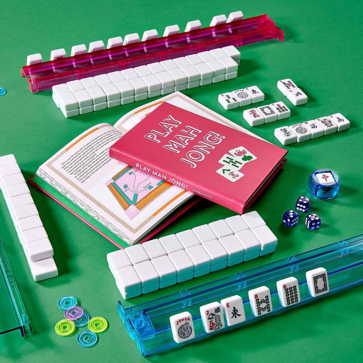 Graphic Image’s Play Mahjong! game set—featuring racks, tiles, dice, tokens, and the open “Play Mahjong!” book—is attractively arranged on a green surface and makes an ideal choice for anyone looking to play mahjong and master winning strategies.