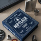 A box labeled "The Essentials Hip Flask" by PrintWorks sits on a table alongside a stainless steel flask, shot glasses, and other items.