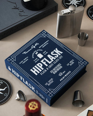 A box labeled "The Essentials Hip Flask" by PrintWorks sits on a table alongside a stainless steel flask, shot glasses, and other items.