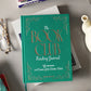 The PrintWorks Book Club Reading Journal, a green hardcover book, rests on a table with glasses, a teapot, and other books—ideal for tracking your reading progress and writing book reviews.