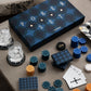 The PrintWorks Art of Poker Set, featuring blue and gold chips, pro-grade playing cards, and two glasses for ice water, sits on a table beside a patterned chair—ideal for mastering poker skills.