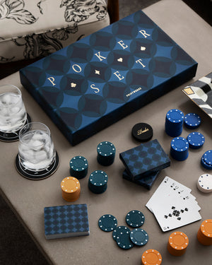 The PrintWorks Art of Poker Set, featuring blue and gold chips, pro-grade playing cards, and two glasses for ice water, sits on a table beside a patterned chair—ideal for mastering poker skills.