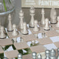 A close-up of the PrintWorks Chess Collection—a beige and silver set on a mirrored board—makes an elegant coffee table game or classic addition to your collection. Books and a vase complete the sophisticated scene.