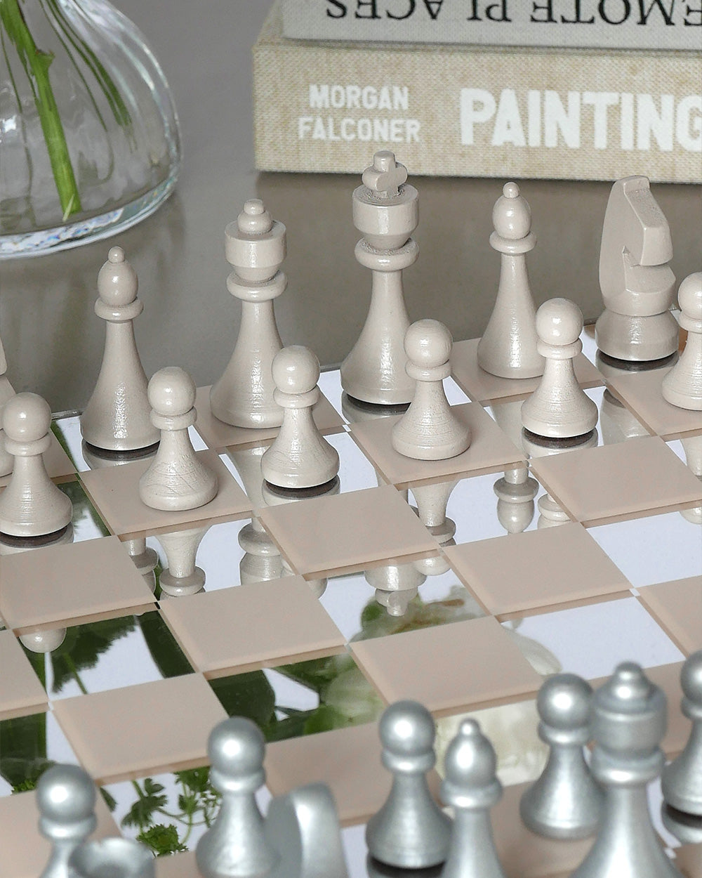 A close-up of the PrintWorks Chess Collection—a beige and silver set on a mirrored board—makes an elegant coffee table game or classic addition to your collection. Books and a vase complete the sophisticated scene.