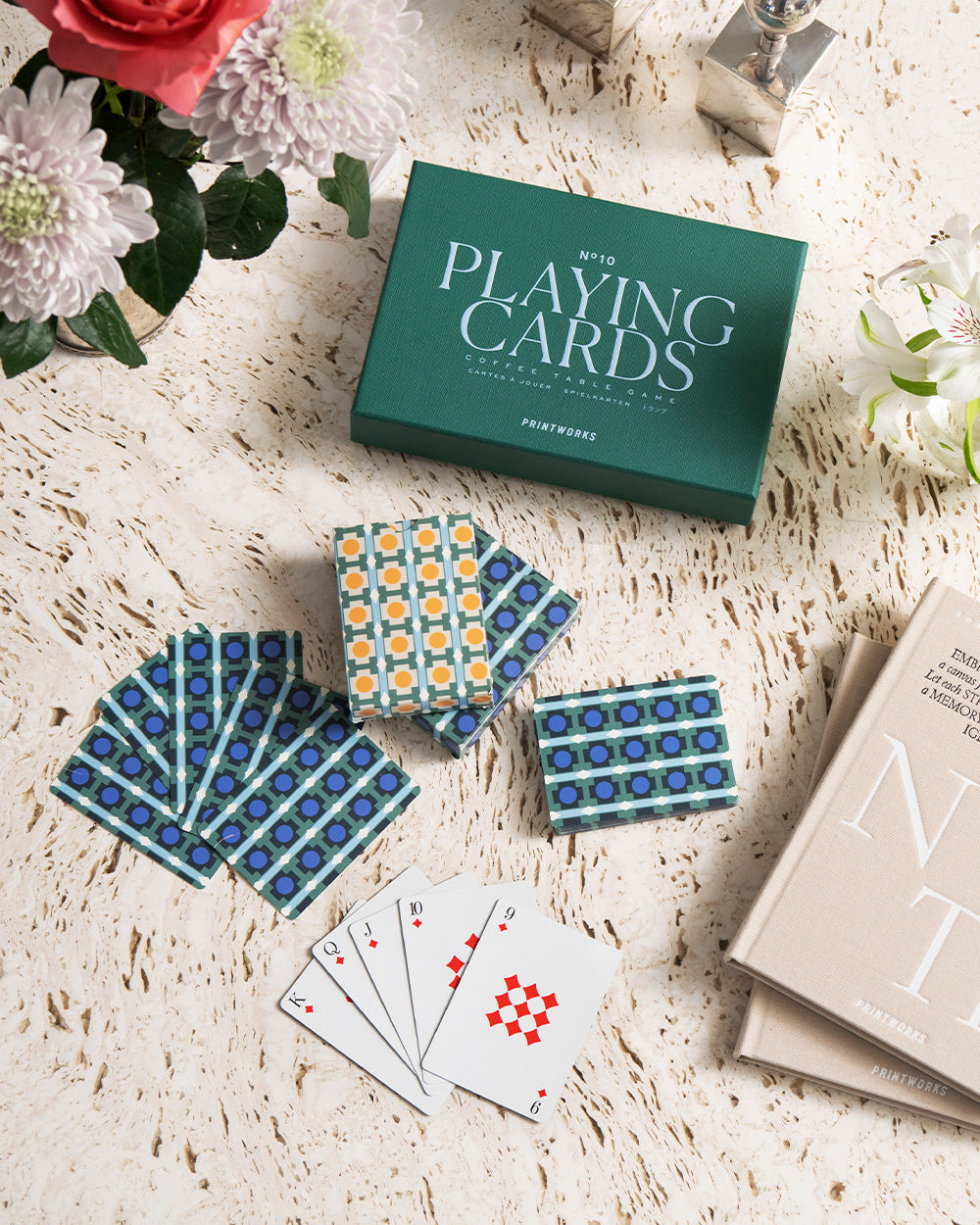 A set of PrintWorks Playing Cards is arranged on a marble table beside a green box, flowers, and beige books. Four cards with red diamonds are face up.