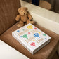 A brown teddy bear sits beside the PrintWorks "Dream Big Little One Photo Album," featuring colorful hot air balloons on the cover, all arranged on a brown upholstered chair—an adorable baby milestone photo display.