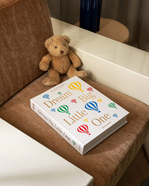 A brown teddy bear sits beside the PrintWorks "Dream Big Little One Photo Album," featuring colorful hot air balloons on the cover, all arranged on a brown upholstered chair—an adorable baby milestone photo display.
