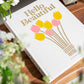 The PrintWorks Hello Beautiful Photo Album features a white cover with yellow and pink tulip illustrations, resting on a wooden surface with flowers—an ideal coffee table piece or stylish home decor accent.