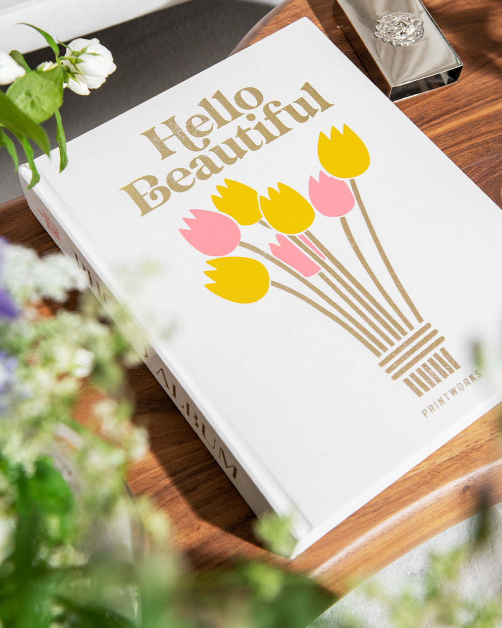 The PrintWorks Hello Beautiful Photo Album features a white cover with yellow and pink tulip illustrations, resting on a wooden surface with flowers—an ideal coffee table piece or stylish home decor accent.