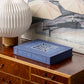 A closed Blue Picture Perfect Photo Album by PrintWorks sits on a wooden dresser next to a white geometric lamp and a framed umbrella art print.