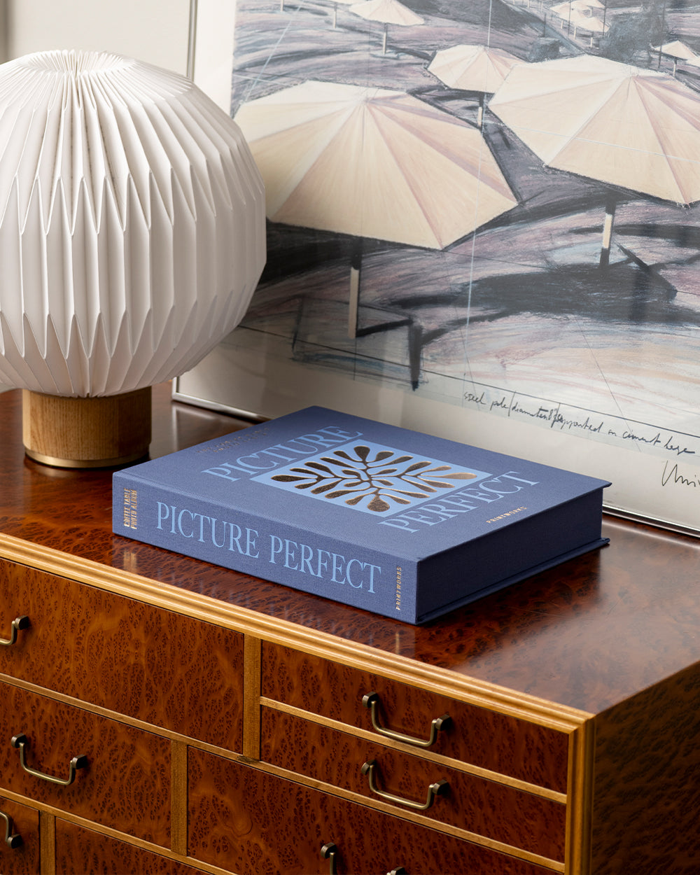 A closed Blue Picture Perfect Photo Album by PrintWorks sits on a wooden dresser next to a white geometric lamp and a framed umbrella art print.