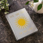The PrintWorks "You Are The Sunshine Of My Life Photo Album" features a silver cover with a yellow sun design and uplifting words, making it an elegant piece for your home decor when displayed on a dark marble table with white flowers.