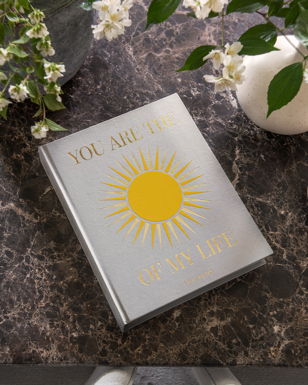 The PrintWorks "You Are The Sunshine Of My Life Photo Album" features a silver cover with a yellow sun design and uplifting words, making it an elegant piece for your home decor when displayed on a dark marble table with white flowers.