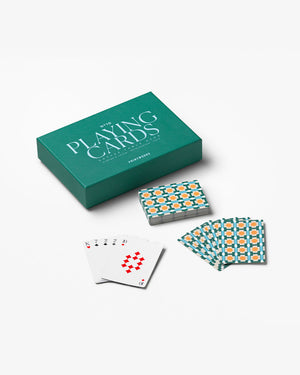 A green PrintWorks "Playing Cards" box sits next to a deck of decorative playing cards, with several cards spread out—some face-up displaying red diamonds and black spades.