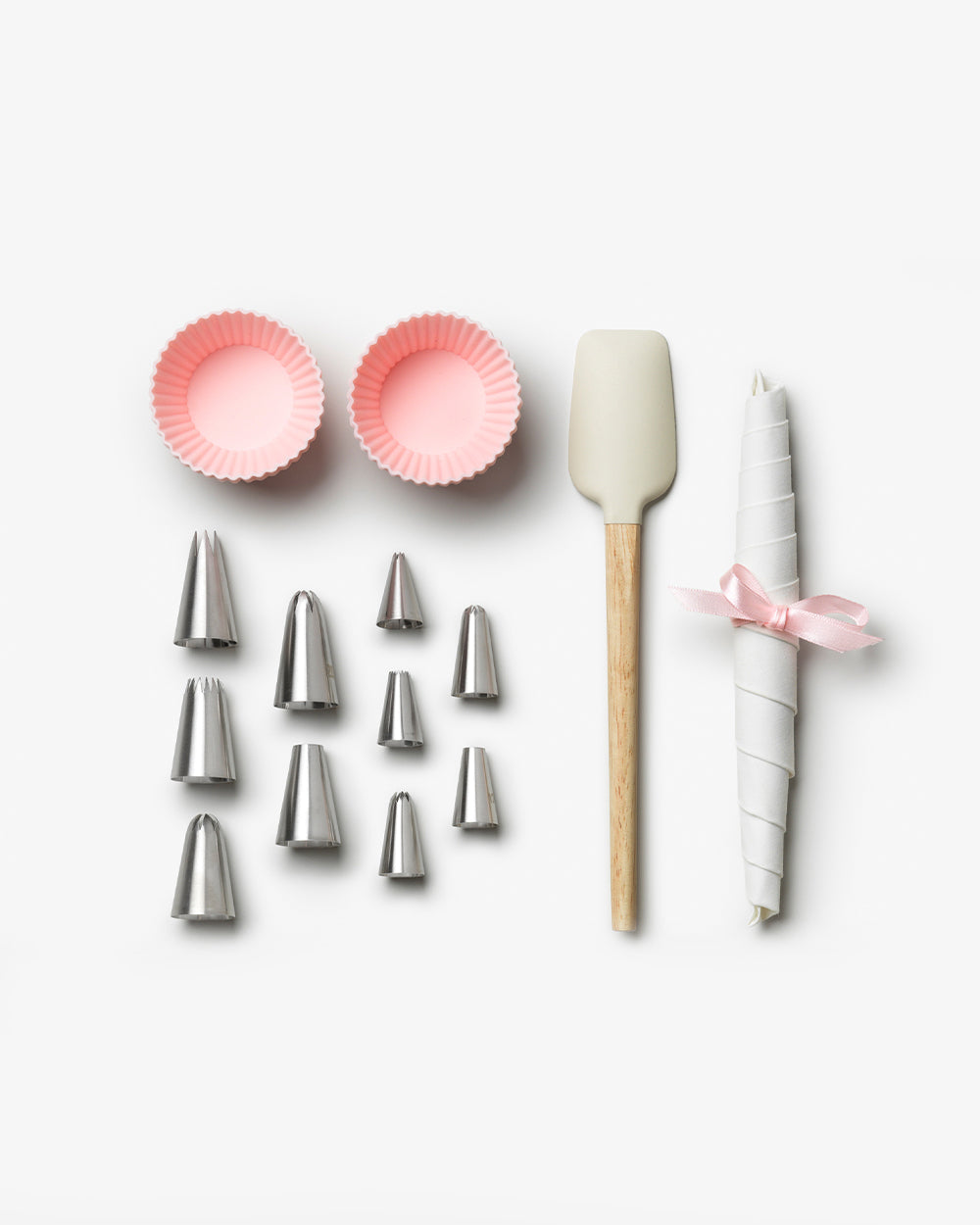 The Essentials Cupcake Tools by PrintWorks includes two pink cupcake liners, nine metal piping tips, a silicone spatula with wooden handle, and rolled parchment tied with a pink ribbon—all presented on a white background.