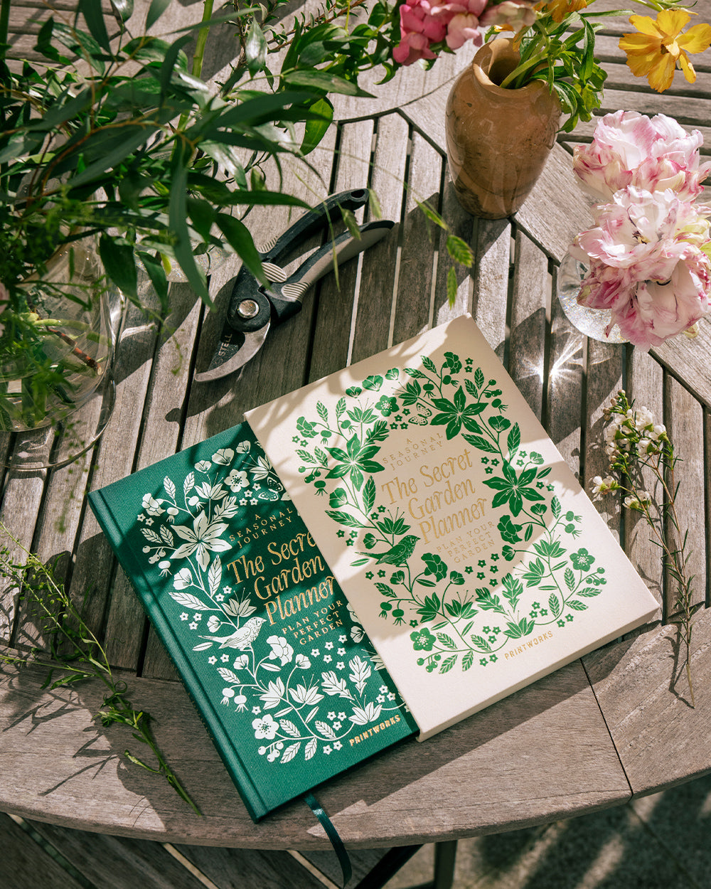 Two hardcover "Secret Garden Journal" books by PrintWorks rest on a round wooden table with garden shears, a vase, assorted flowers, and room for your gardening journal or plant tracker notes.