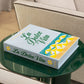 A PrintWorks La Dolce Vita Photo Album sits atop a round green glass coffee table placed between two beige upholstered chairs.