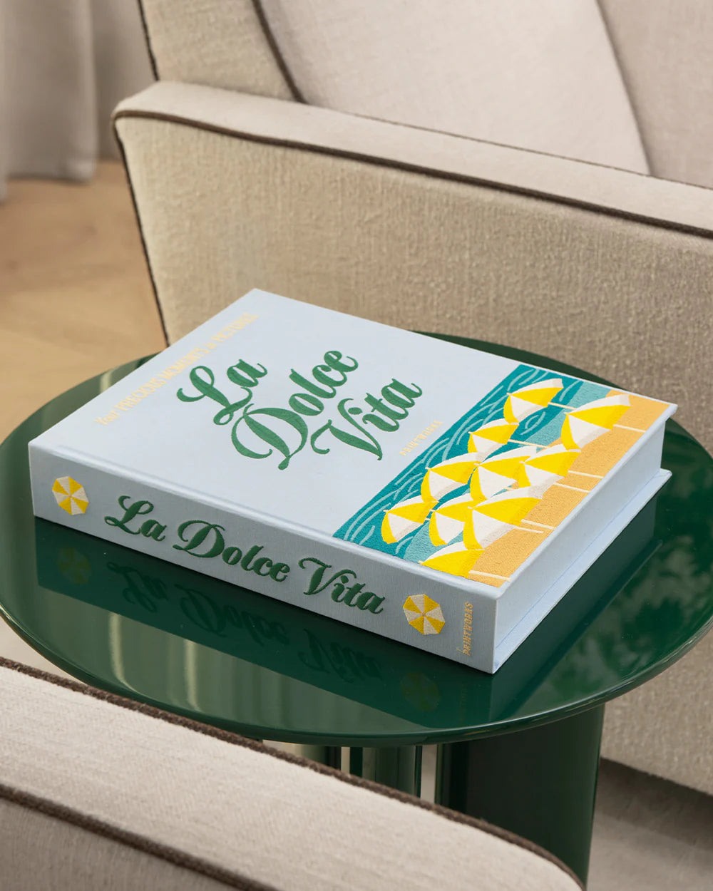 A PrintWorks La Dolce Vita Photo Album sits atop a round green glass coffee table placed between two beige upholstered chairs.