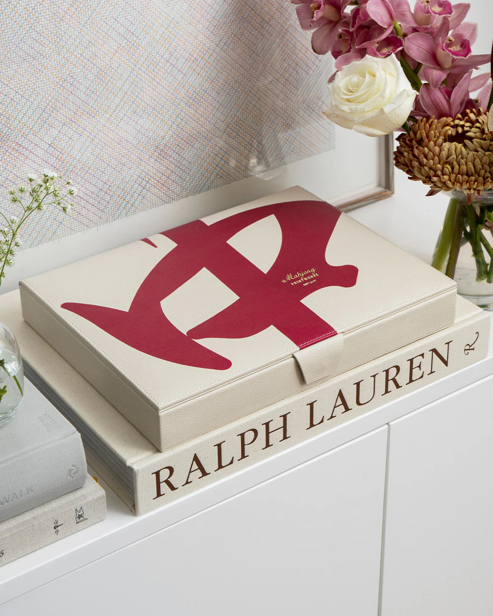 The PrintWorks Mahjong Reverra Game Set, featuring a bold red abstract design, rests on a Ralph Lauren book and is surrounded by vases of flowers atop a white cabinet.