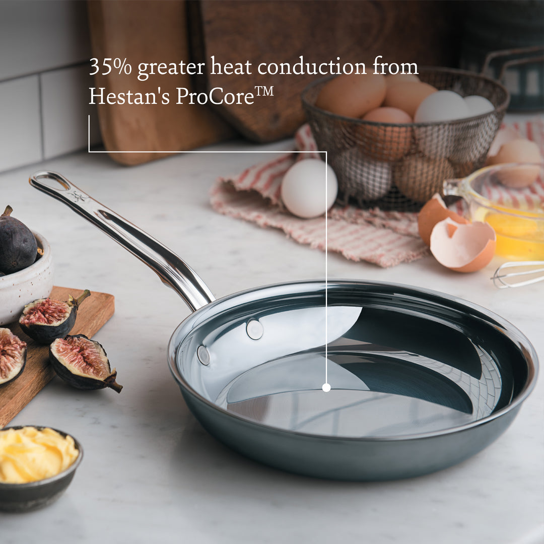 Hestan's NanoBond Titanium Ultimate Cookware 10-Piece Set on a kitchen counter with food items; text reads “35% greater heat conduction from Hestan's ProCore aluminum.”.