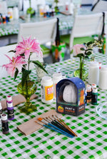 A table featuring Hive’s Paint a Ceramic Pumpkin Event kit—complete with paint supplies, brushes, flowers in vases, and set atop a green and white checkered tablecloth—is perfect for a fun kids’ Halloween activity or for hosting your own ceramic pumpkin painting class in West Palm Beach.