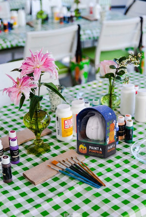 A table featuring Hive’s Paint a Ceramic Pumpkin Event kit—complete with paint supplies, brushes, flowers in vases, and set atop a green and white checkered tablecloth—is perfect for a fun kids’ Halloween activity or for hosting your own ceramic pumpkin painting class in West Palm Beach.