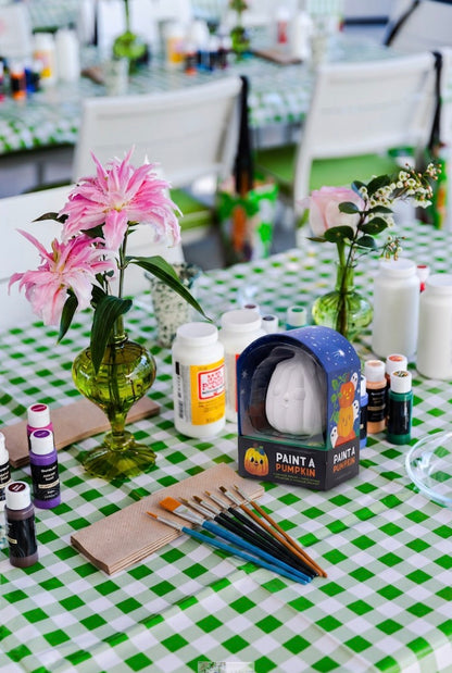 A table featuring Hive’s Paint a Ceramic Pumpkin Event kit—complete with paint supplies, brushes, flowers in vases, and set atop a green and white checkered tablecloth—is perfect for a fun kids’ Halloween activity or for hosting your own ceramic pumpkin painting class in West Palm Beach.